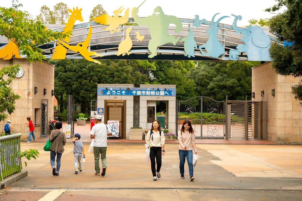 A park entrance with a large sign featuring animal silhouettes and a blue building with Japanese characters.