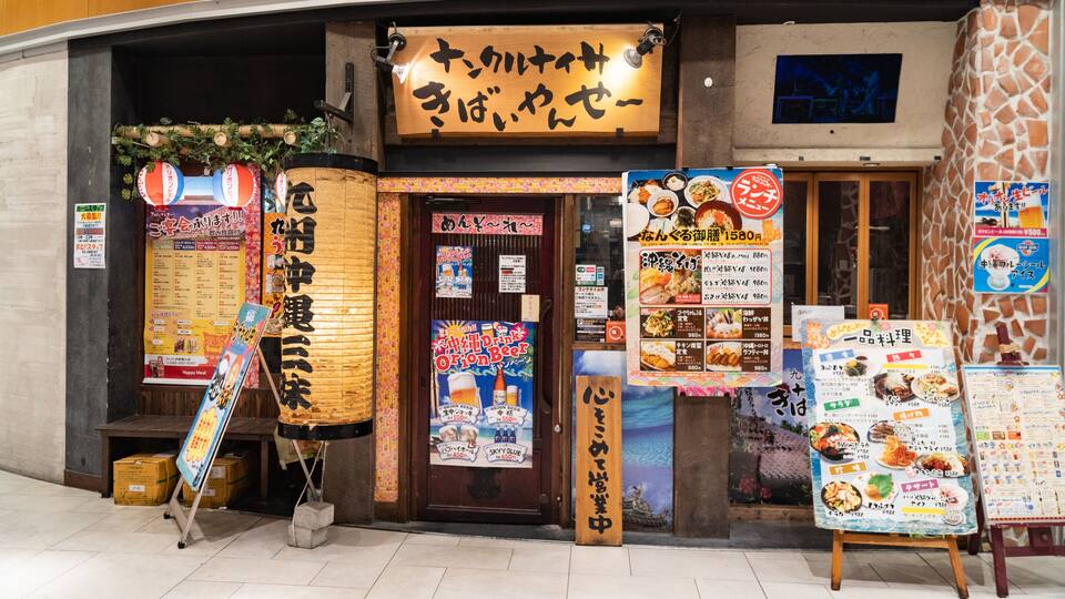 A Japanese restaurant with a traditional wooden exterior, a large signboard, and various posters advertising food and drinks.
