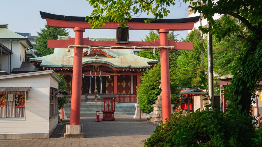 A traditional Japanese torii gate with a green and red roof, surrounded by lush greenery and modern buildings.