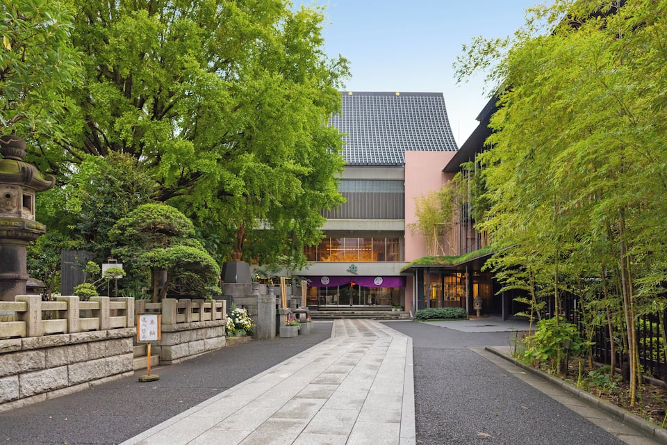 Ein traditionelles japanisches Gebäude mit moderner Glasfassade, umgeben von üppigem Grün und einem Kiesweg.