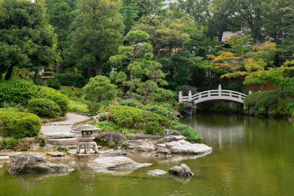 A tranquil Japanese garden with a stone bridge over a pond, surrounded by lush greenery and traditional stone lanterns.