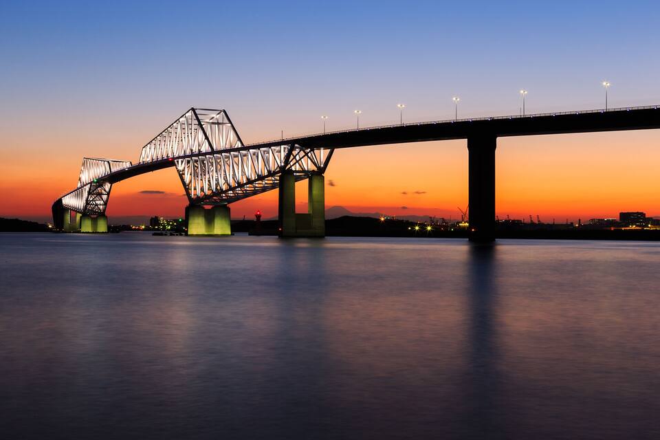 Uma ponte treliçada de aço ao entardecer, com luzes acesas, atravessando um corpo de água.
