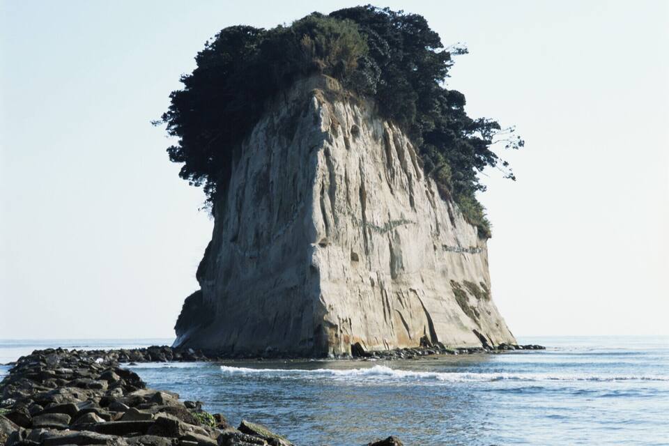 A large rock formation with a steep cliff and a small cave at its base, surrounded by calm sea water and rocky shoreline.