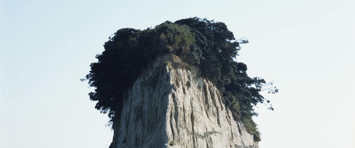 A large rock formation with a steep cliff and a small cave at its base, surrounded by calm sea water and rocky shoreline.