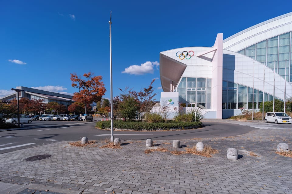 Un bâtiment moderne orné du logo des anneaux olympiques, une place pavée avec des bornes en pierre et des arbres aux couleurs automnales.