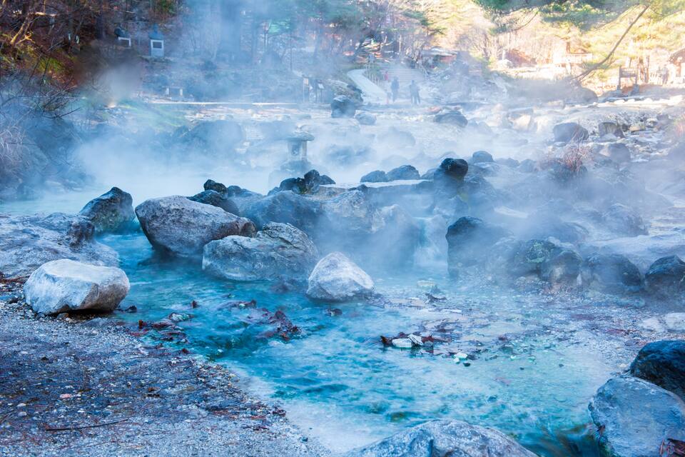 Uma nascente termal geotérmica com vapor subindo da água, rodeada por pedras e árvores.