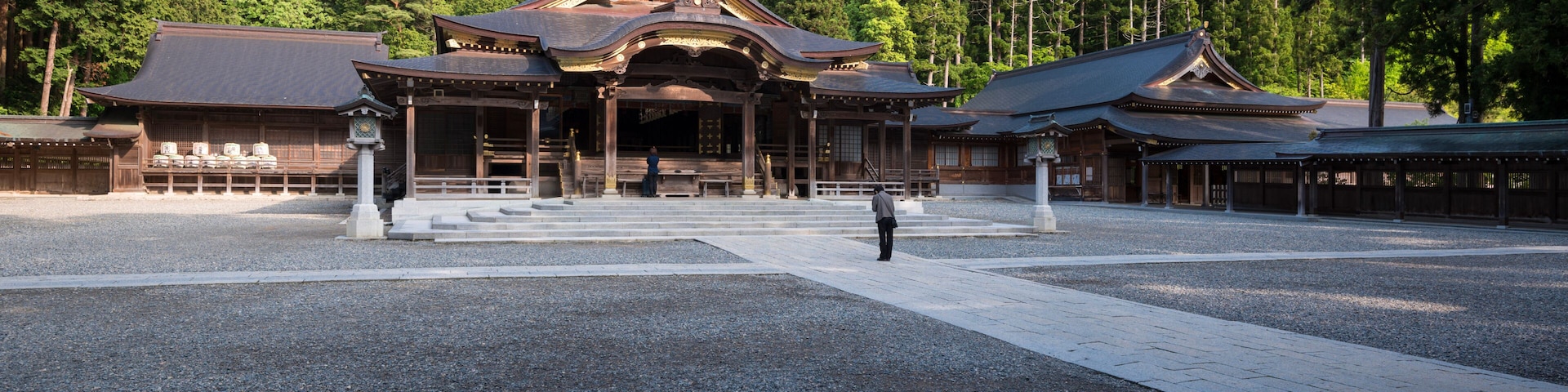 Um templo japonês tradicional com um salão central amplo, rodeado por um pátio de cascalho e colinas arborizadas.