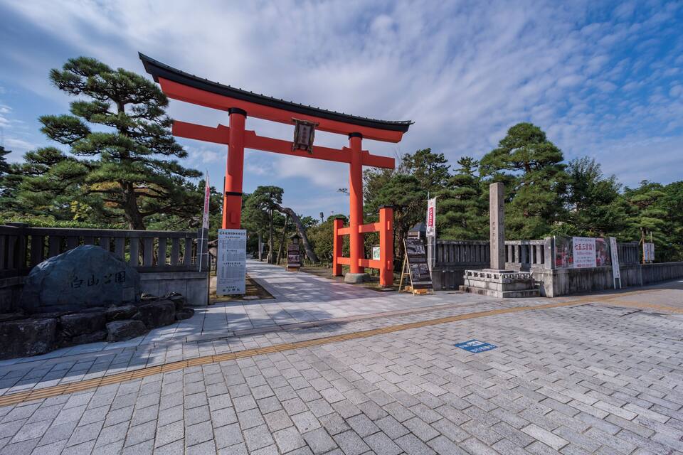 Un torii japonés tradicional con estructura de color rojo y base de piedra, rodeado de árboles y un camino pavimentado.