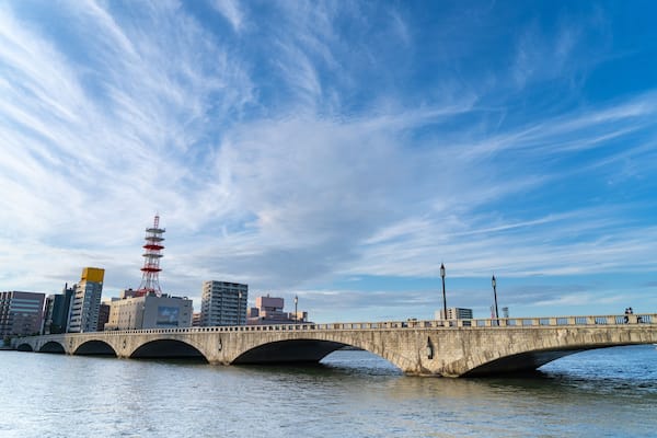 Un pont de pierre enjambant une étendue d’eau, avec une vue sur les bâtiments d’une ville en arrière-plan, dont une tour rouge.