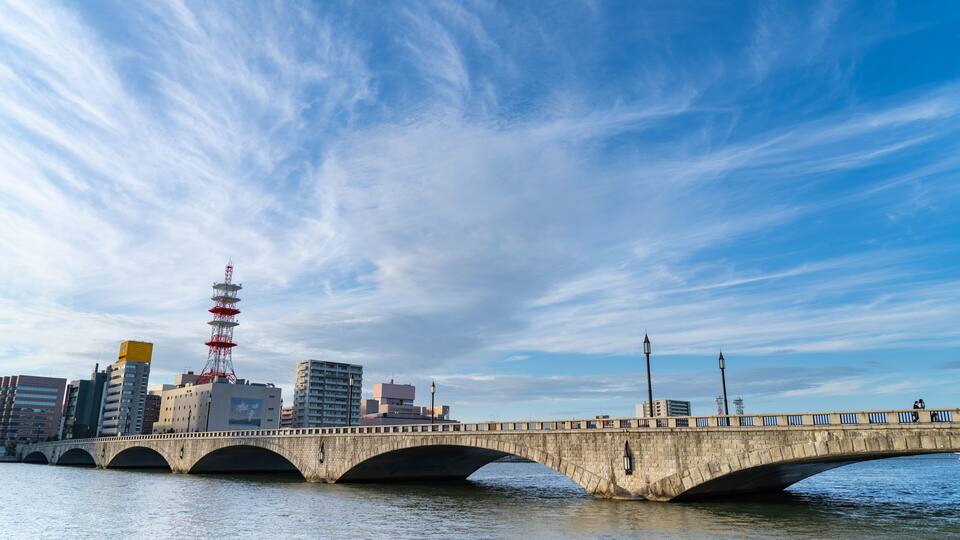 A stone bridge over water with a city skyline in the background, including a red tower.