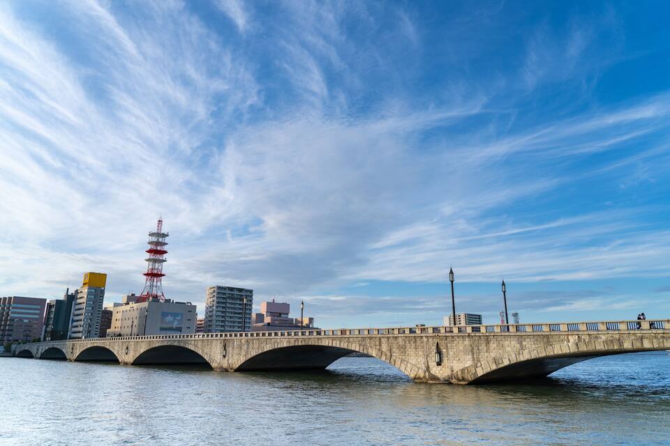 A stone bridge over water with a city skyline in the background, including a red tower.