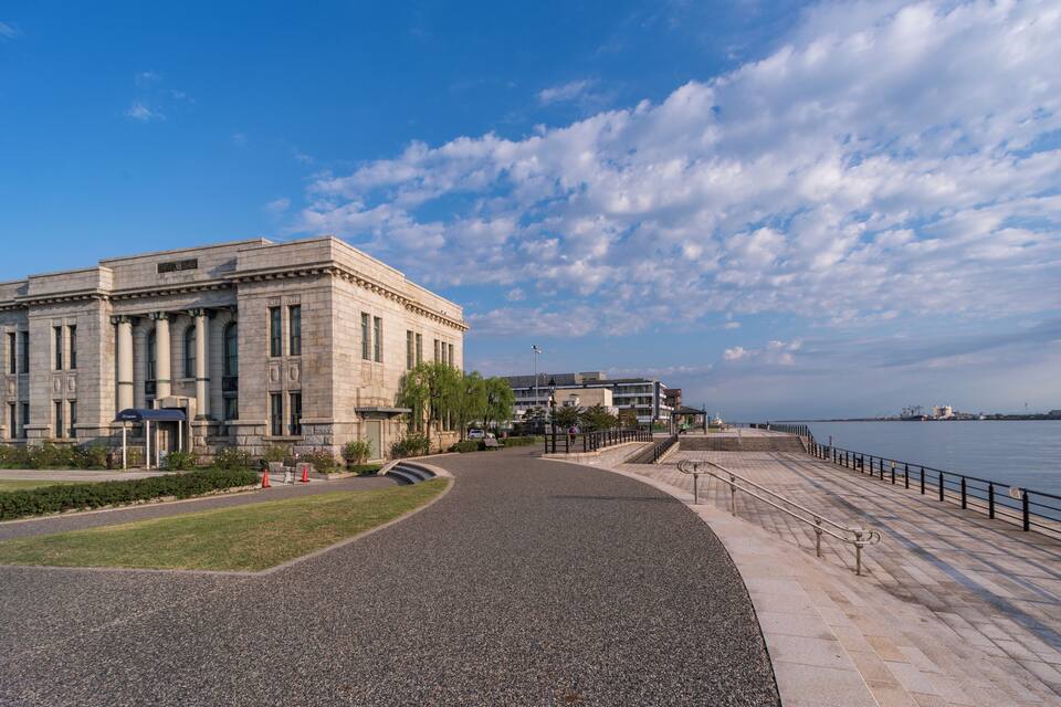 A large classical building with columns, a paved walkway, and a waterfront.