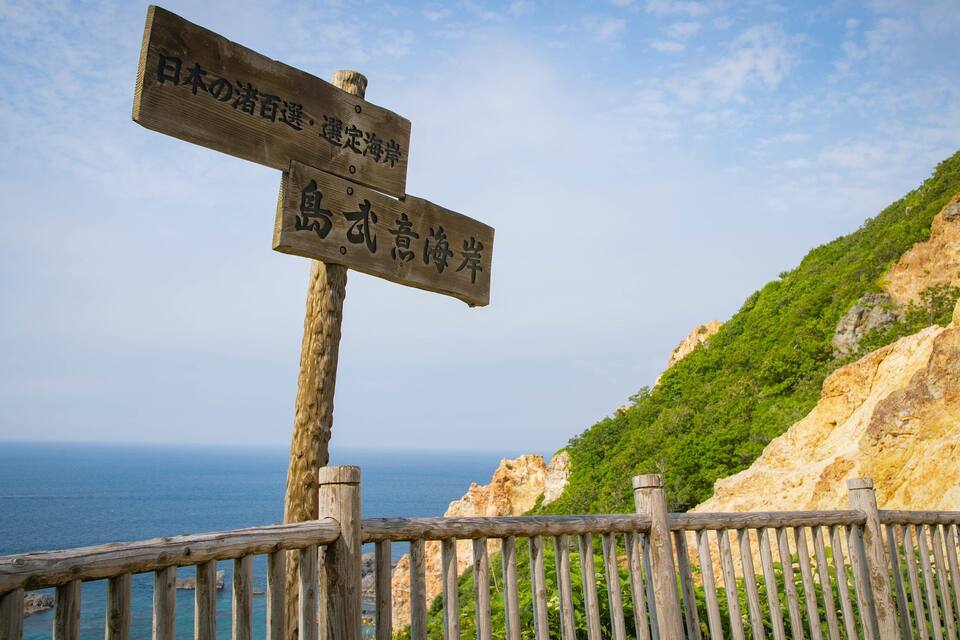 A wooden signpost with Japanese characters, a wooden fence, and a coastal landscape with cliffs and the sea.