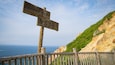 Ein hölzernes Wegweiser mit japanischen Schriftzeichen, ein Holzzaun und eine Küstenlandschaft mit Klippen und dem Meer.