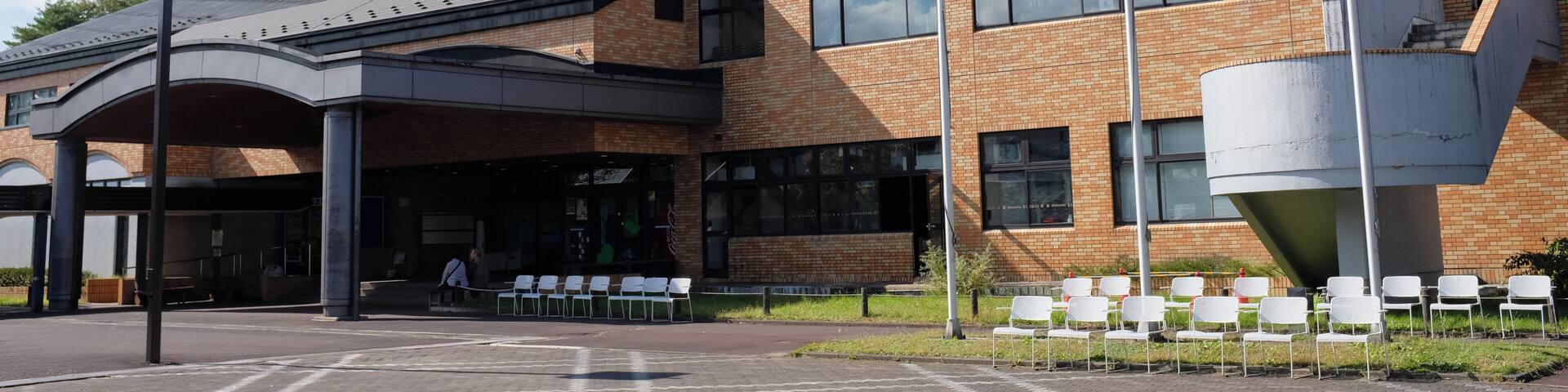 A modern building with a brick facade, large windows, and a covered entrance area with white chairs.
