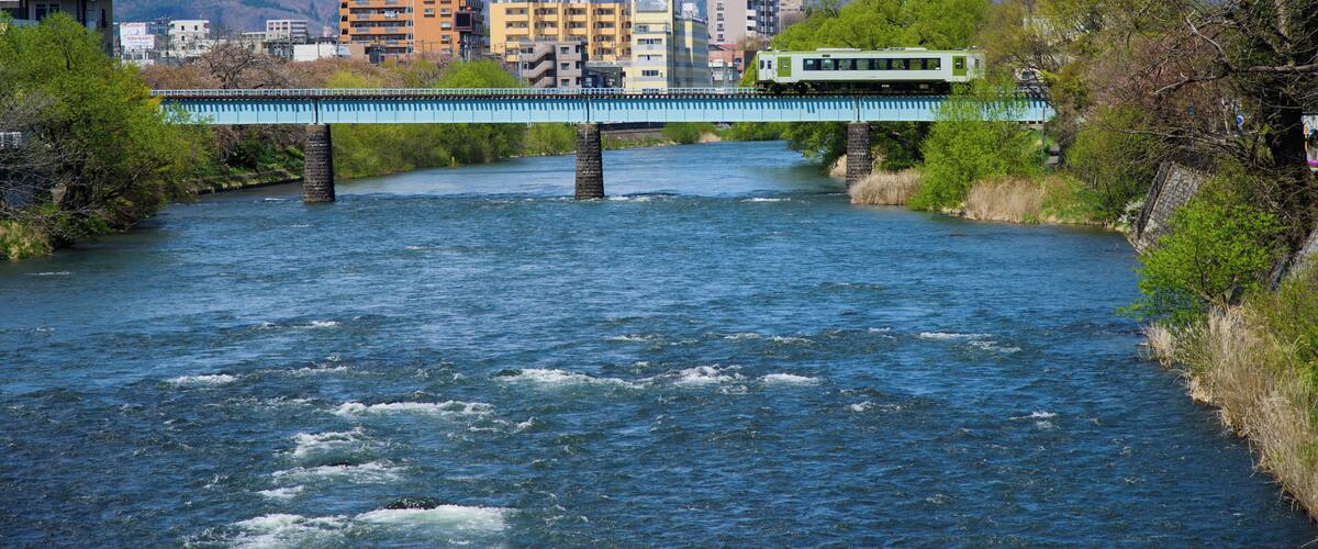A river with a bridge, a green train, and a snow-capped mountain in the background.