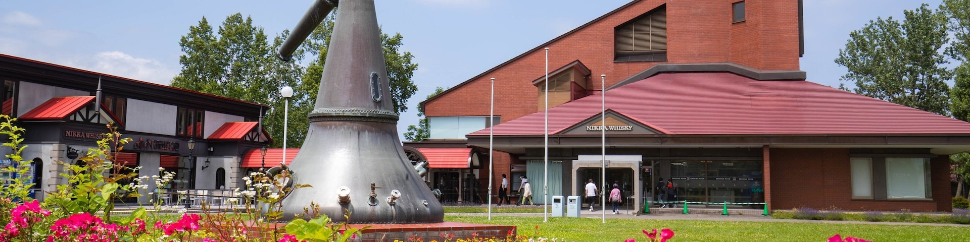 Uma grande escultura metálica em um parque, com um edifício com a inscrição "NIKKA WHISKY" ao fundo.