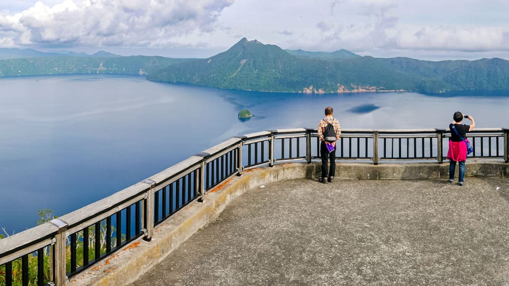Ein weiter Blick auf einen See, umgeben von Bergen, eine hölzerne Aussichtsplattform und eine entfernte Insel.