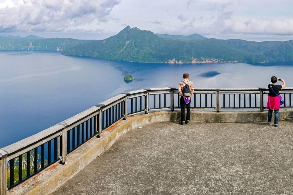 Vista panoràmica d'un llac envoltat de muntanyes, una passarel·la de fusta i una illa a la distància.