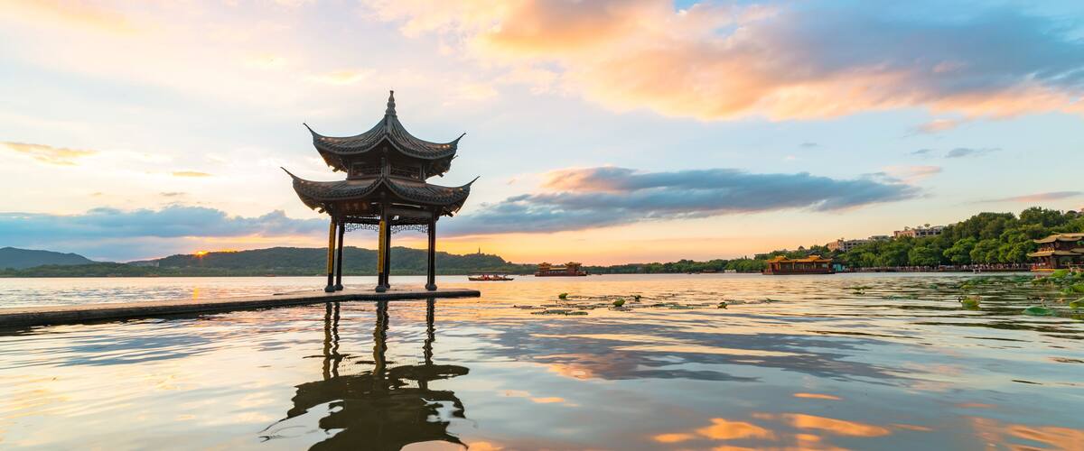 A traditional Chinese pavilion by a calm lake with a sunset sky.
