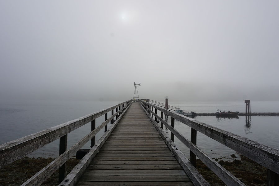 Een houten steiger die zich uitstrekt in een mistig wateroppervlak, met een vuurtoren in de verte.