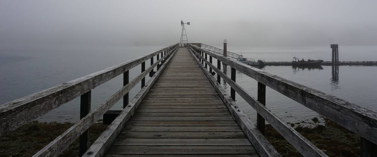 A wooden pier extending into a foggy body of water with a lighthouse in the distance.