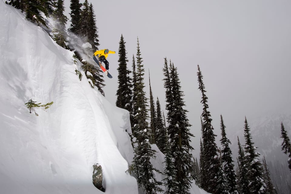 A skier in a yellow jacket performing a jump on a snowy mountain slope with tall evergreen trees.