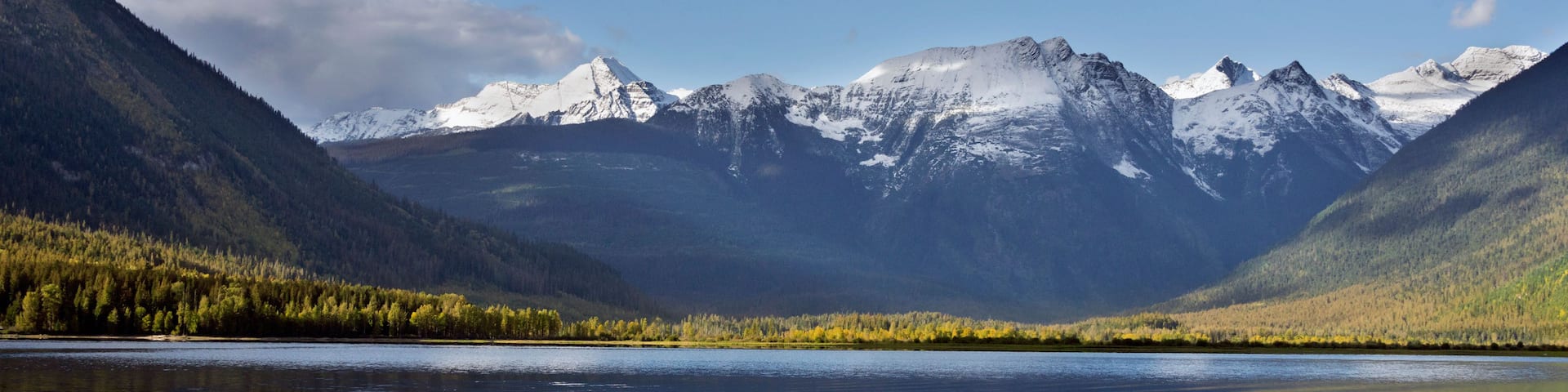 A mountain range with snow-capped peaks, a calm lake reflecting the sky, and dense forested areas.