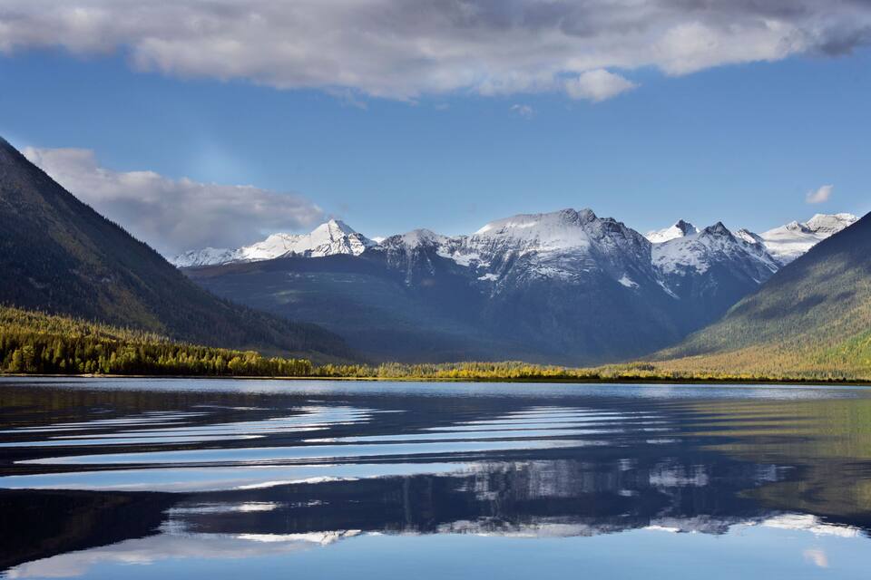 A mountain range with snow-capped peaks, a calm lake reflecting the sky, and dense forested areas.