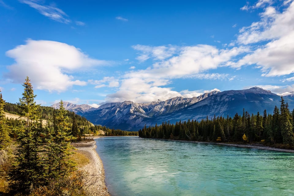 A river with clear turquoise water, surrounded by dense coniferous forests and snow-capped mountains under a partly cloudy sky.