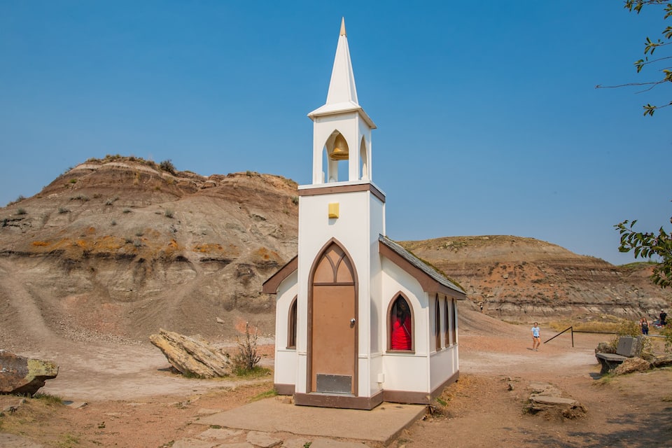 A small white church with a pointed steeple and a red door, surrounded by rocky terrain and sparse vegetation.