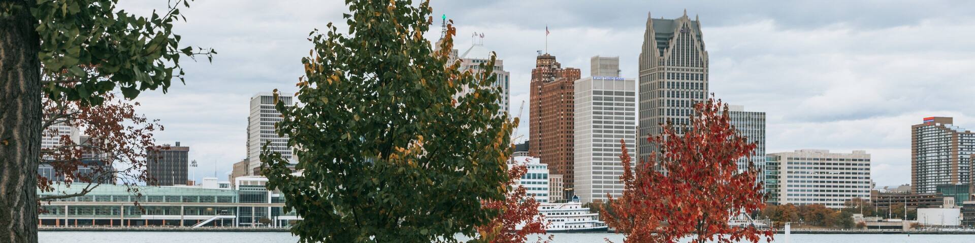 A park with a river, autumn trees, a statue, and a city skyline in the background.