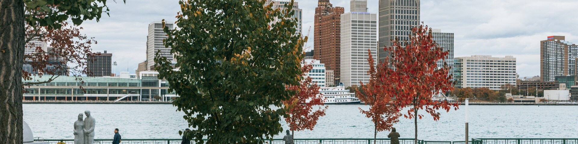 A park with a river, autumn trees, a statue, and a city skyline in the background.