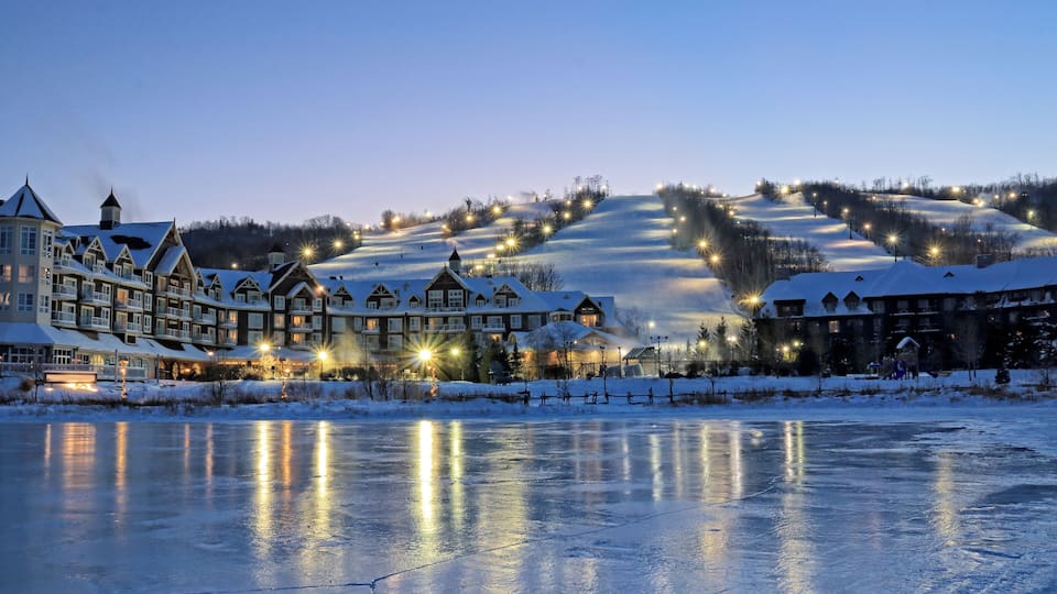 A snow-covered landscape with illuminated buildings and a frozen lake at dusk.