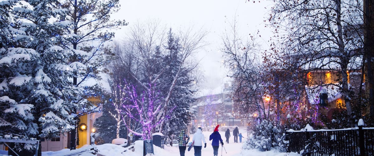 A snowy street with people walking, decorated trees, and buildings in the background.