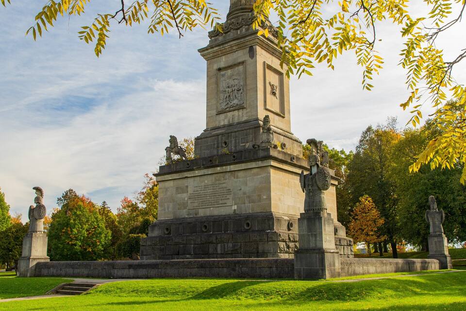 Un monument de pierre orné de statues et d’inscriptions, entouré d’arbres et d’herbe.