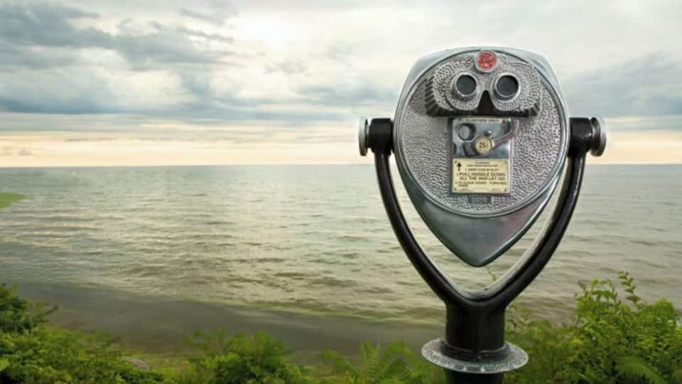 A vintage telescope with a plaque, overlooking a body of water and a cloudy sky.