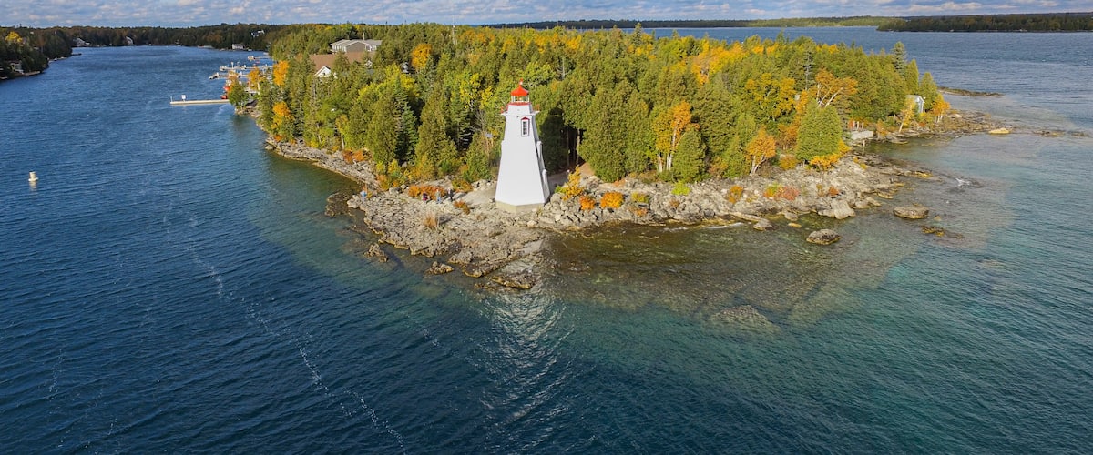 A lighthouse on a rocky island surrounded by clear water and autumn-colored trees.