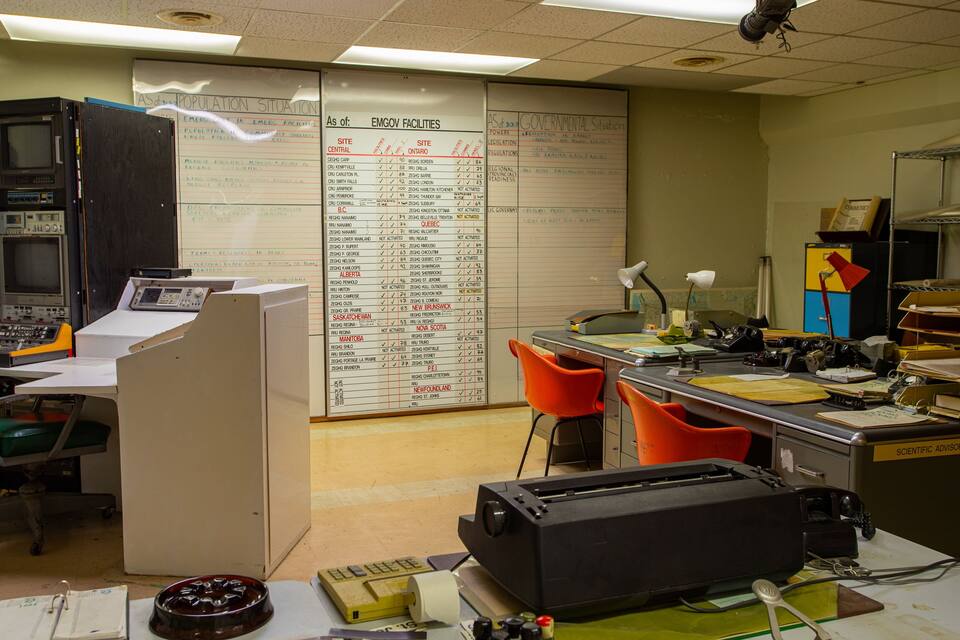 A cluttered office with a whiteboard listing EMGov facilities and a computer with multiple monitors.