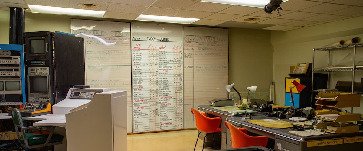 A cluttered office with a whiteboard listing EMGov facilities and a computer with multiple monitors.