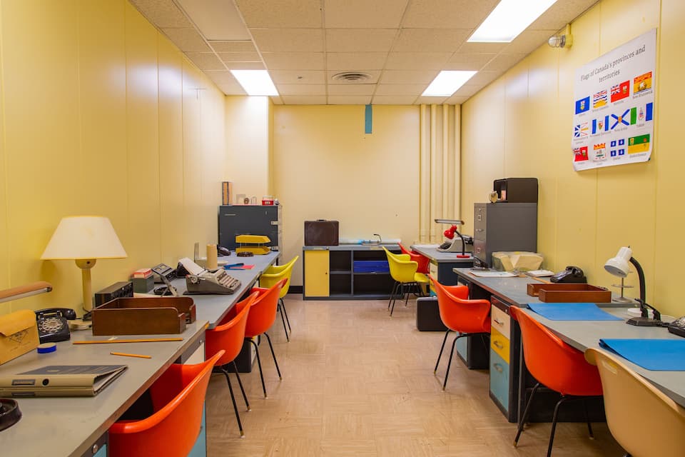 Un aula con paredes amarillas, sillas anaranjadas y azules, escritorios con computadoras y un cartel con banderas canadienses.