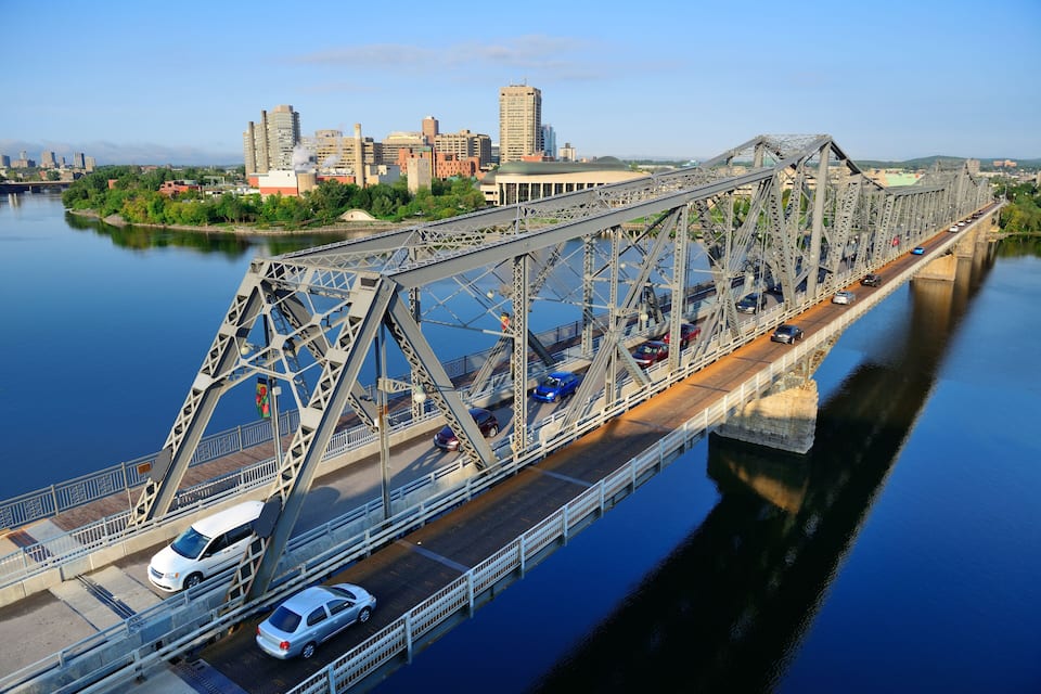 A steel bridge over a calm river with vehicles, a city skyline in the background, and a clear sky.