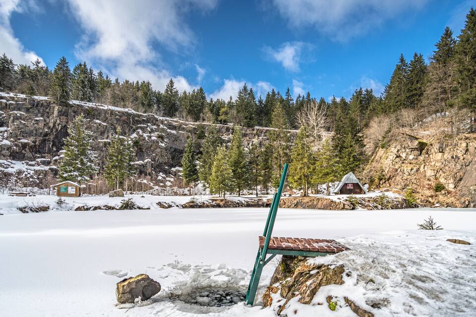 A snow-covered landscape with a frozen lake, a wooden bench, and a small cabin.