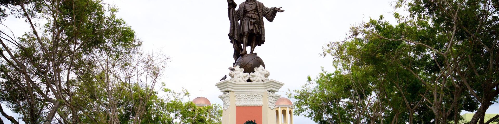 A statue with a fountain in front of a building with a wreath on the pillar.