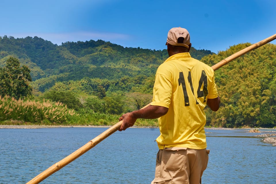 A person in a yellow shirt with the number 14 paddling a canoe on a river surrounded by lush green hills.
