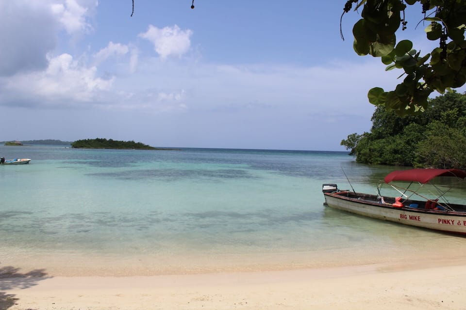 Una playa con agua turquesa cristalina, un barco llamado 'BIG MIKE' con toldo rojo, y una pequeña isla a lo lejos.