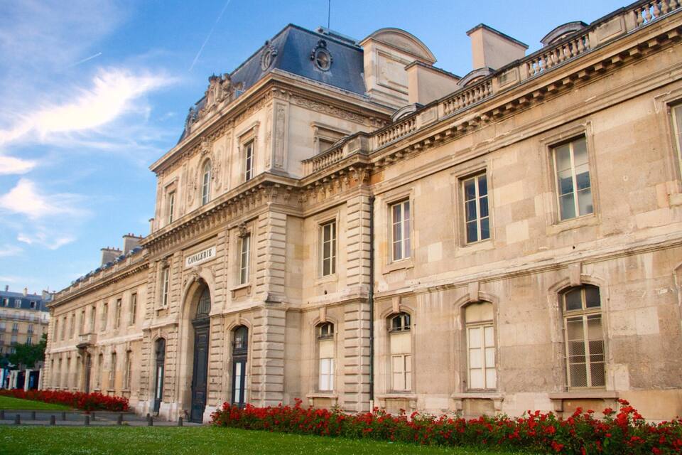 A historic building with classical architecture, a prominent entrance, and a well-maintained garden with red flowers.