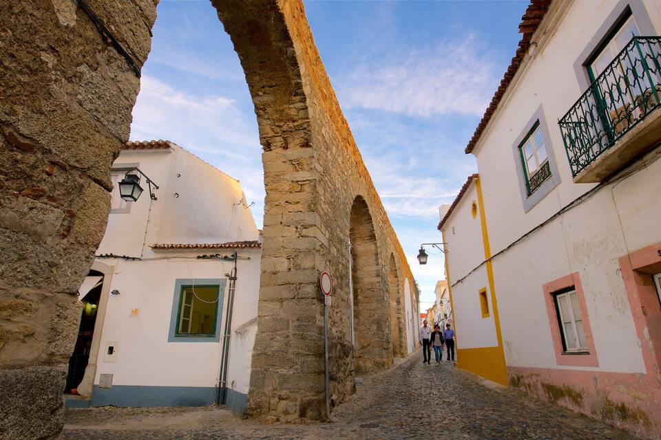 A cobblestone street flanked by historic stone buildings with arched doorways and a clear blue sky.