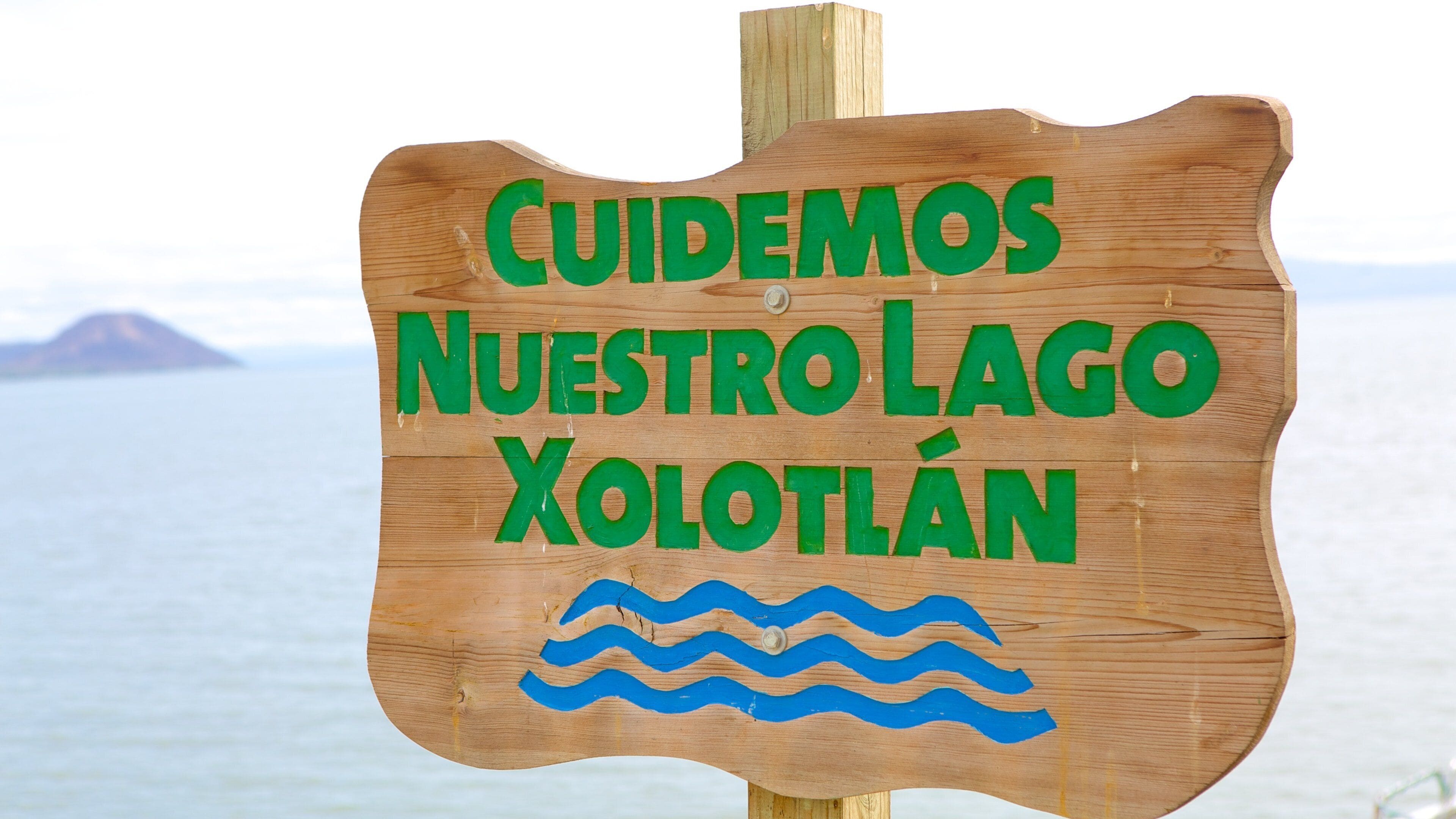 Une pancarte en bois avec le texte vert suivant : « CUIDEMOS NUESTRO LAGO XOLOTLAN » et des motifs bleus représentant des vagues.