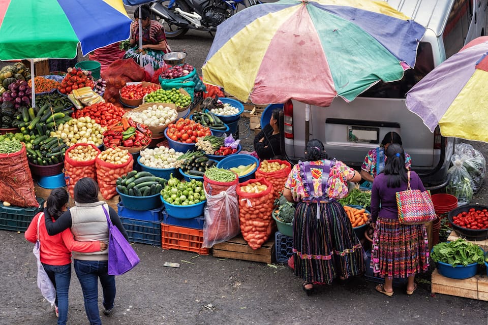 A vibrant outdoor market with various vegetables displayed in large bins and baskets, and people shopping.